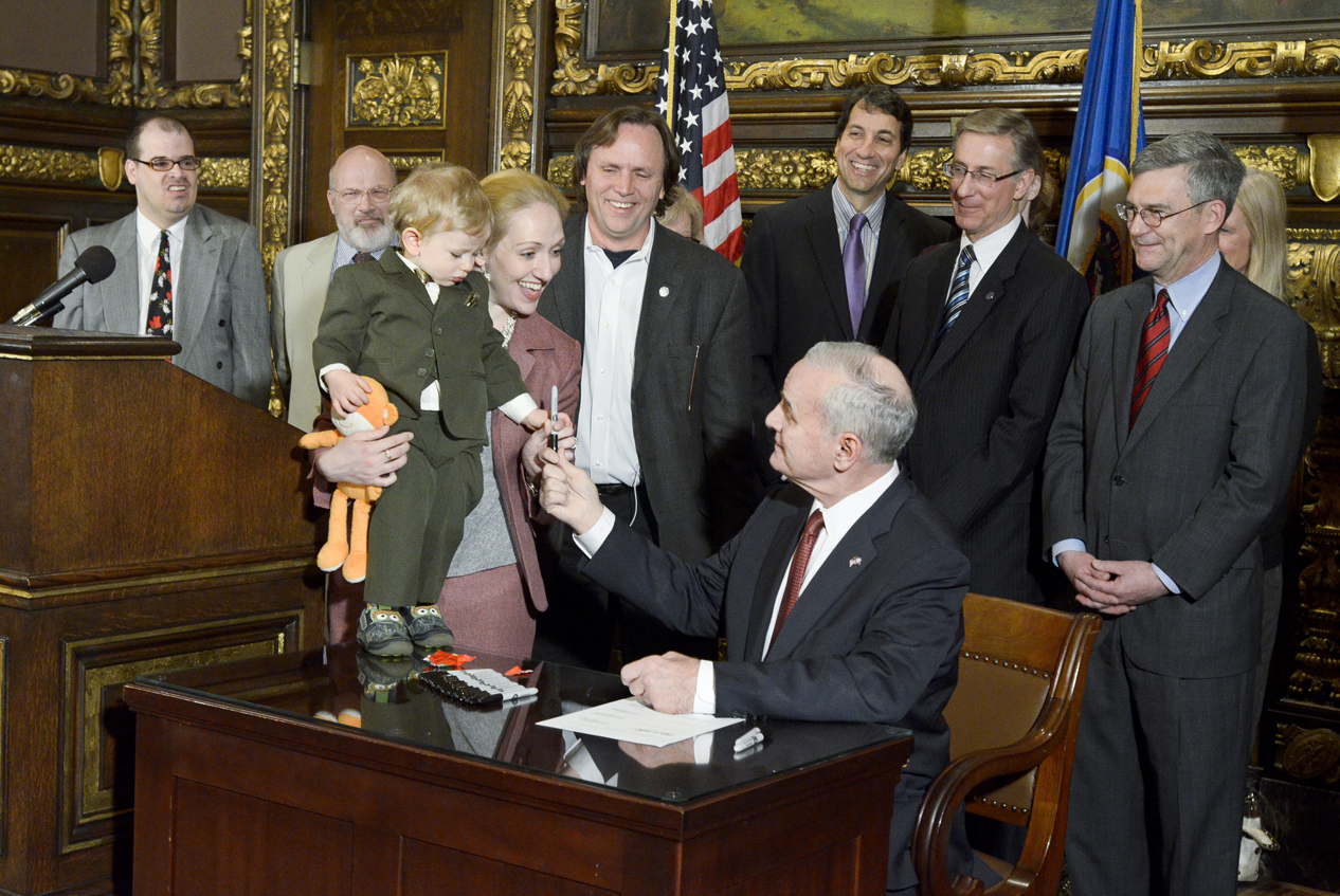 Surrounded by legislators and interested parties, Gov. Mark Dayton presents Alexandra Fitzsimmons, legislative affairs and advocacy director for Children�s Defense Fund-Minnesota, and her 20-month-old, Frankie, a pen used to sign the omnibus health and human services law during an April 30 ceremony. (Photo by Andrew VonBank)